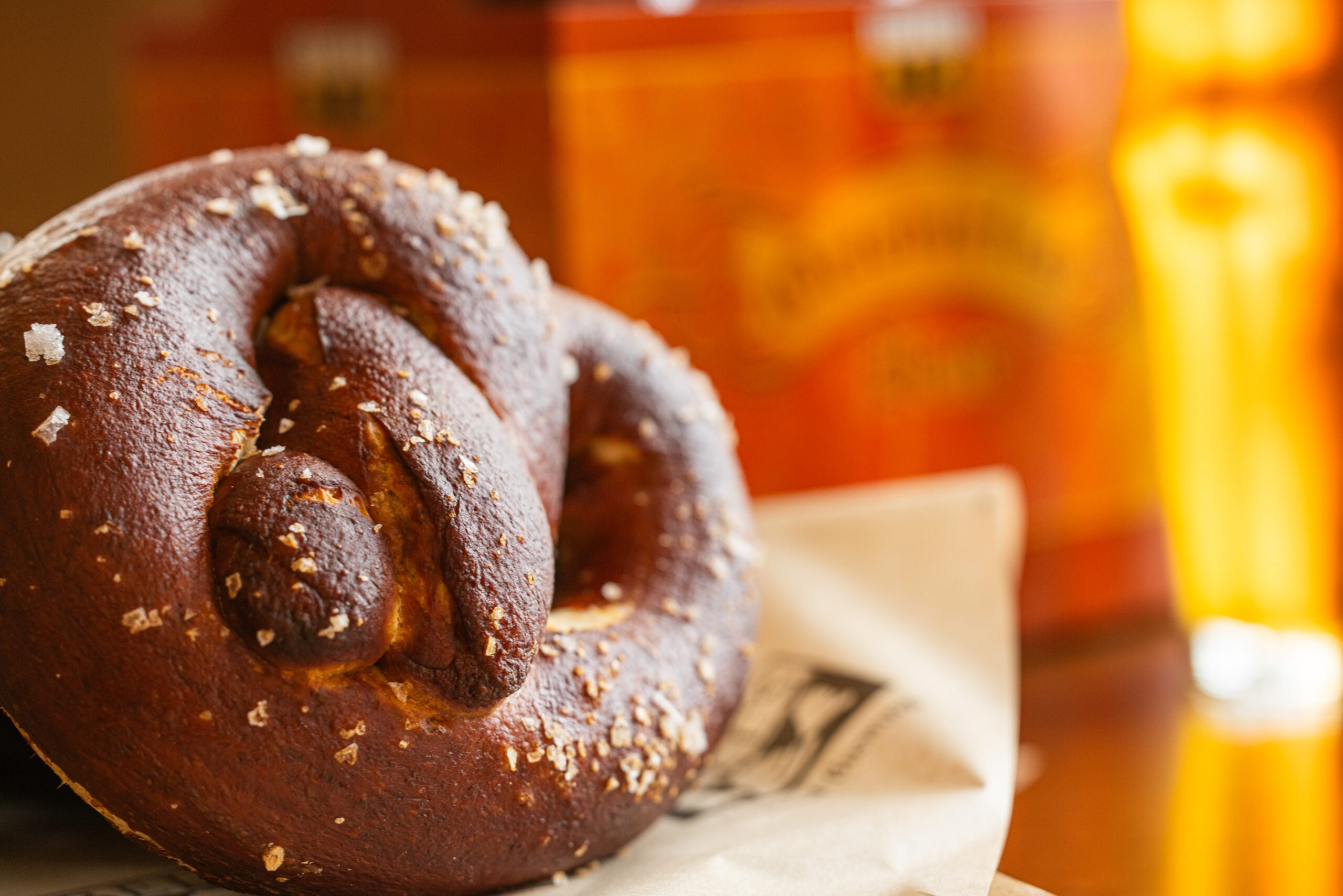 A pretzel in the foreground with a blurred pint of amber colored beer and a six pack of Octoberfest