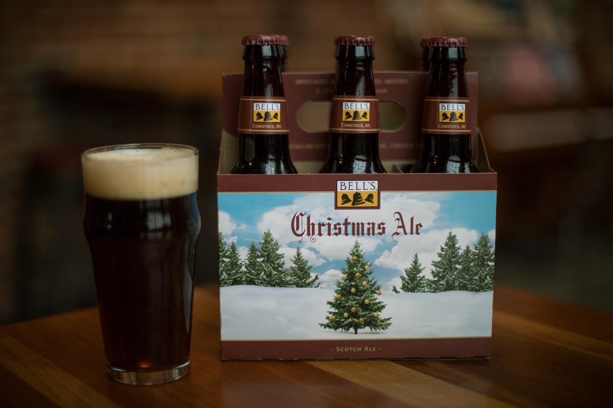 A six-pack of Bell's Christmas Ale positioned on a wooden table, with one beer poured into a clear pint glass showing a rich, dark color and a frothy head. The packaging of the beer features a serene winter landscape with a prominently displayed Christmas tree and the Bell's Brewery logo at the top.