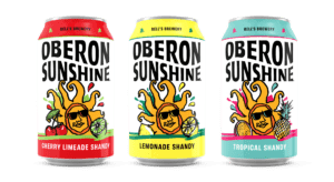 "Three cans of 'Oberon Sunshine' shandy from Bell's Brewery, each with a unique flavor. From left to right: 'Cherry Limeade Shandy' with a red tab and cherries and a lime depicted, 'Lemonade Shandy' with a yellow tab and a lemon slice, and 'Tropical Shandy' with a teal tab and a pineapple and orange slice. Each can has a sun with sunglasses in the center, varying background colors, and the Bell's Brewery logo at the top."